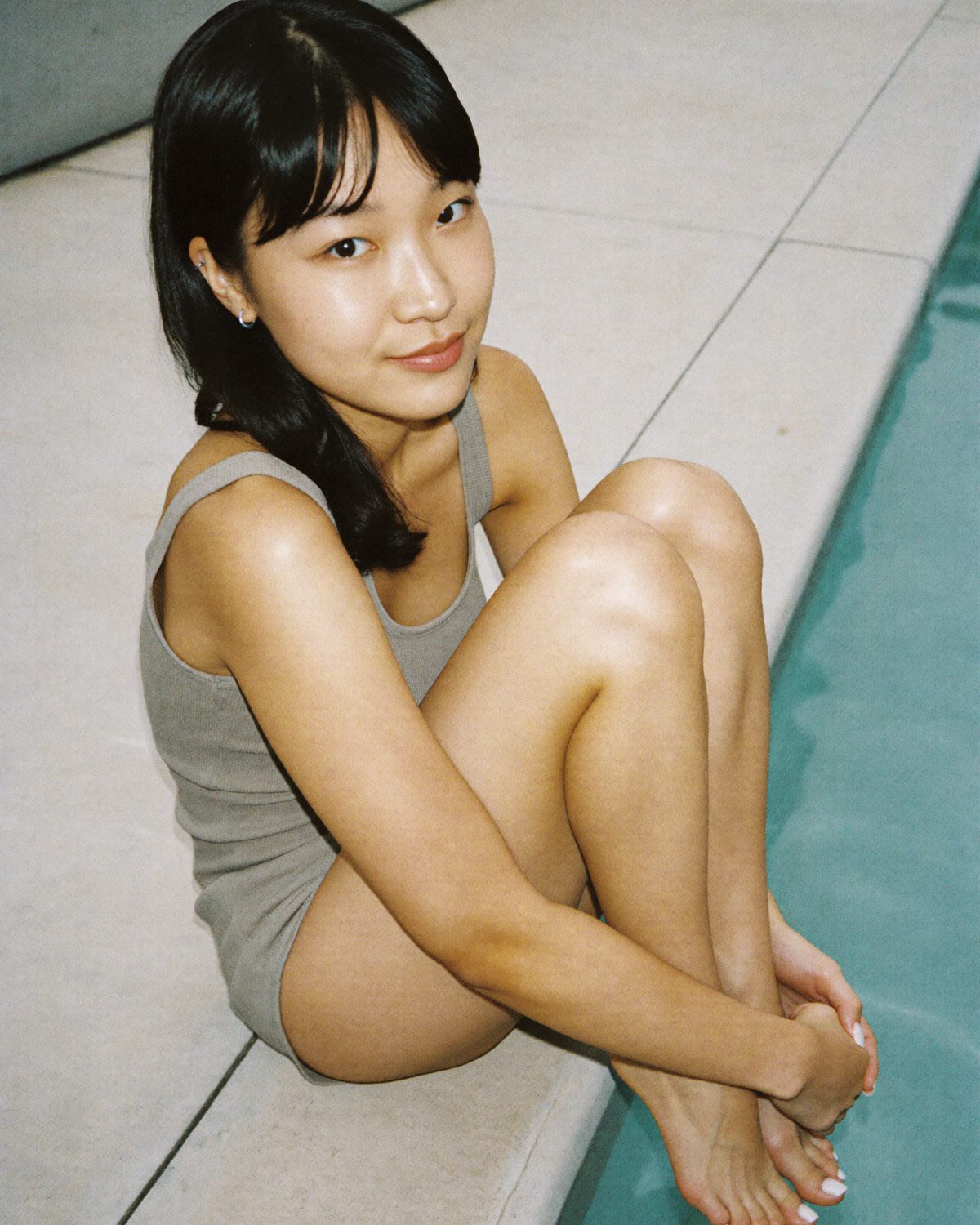 A young woman with dark hair sitting at the edge of a pool with her knees pulled up, bare feet with white polish
