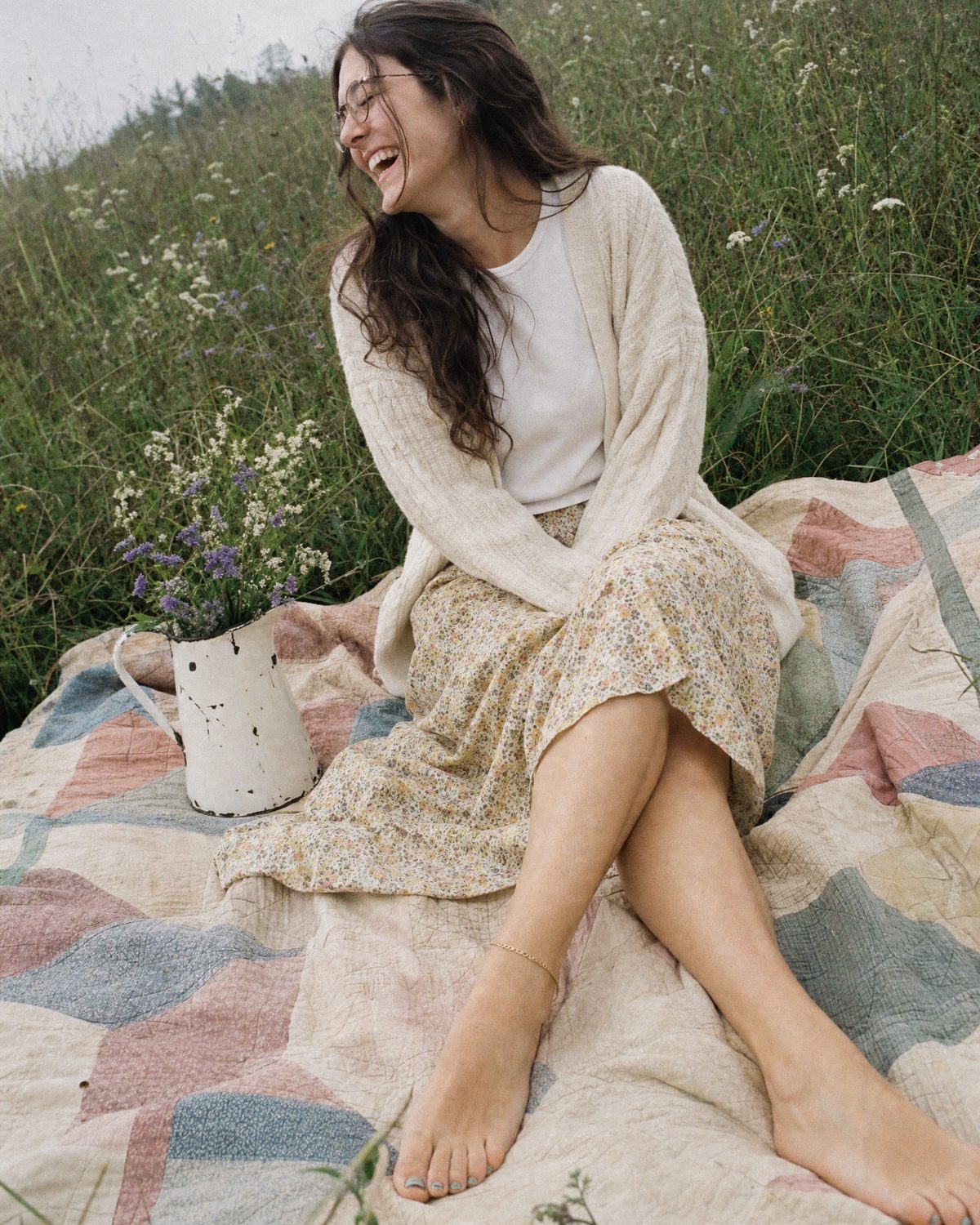 A young woman in glasses laughing on a faded quilt in a wildflower meadow, bare feet with a thin gold anklet