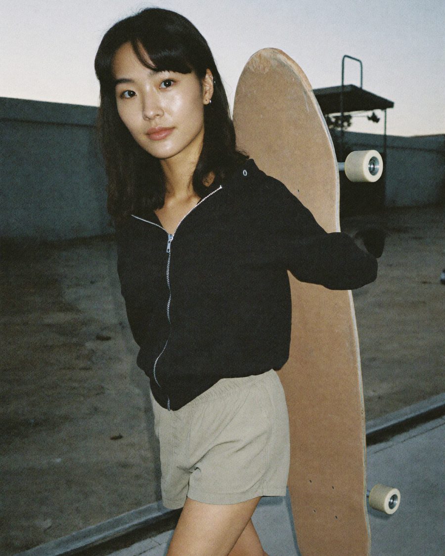 A young woman with dark hair carrying a longboard over her shoulder at dusk, black zip hoodie and khaki shorts