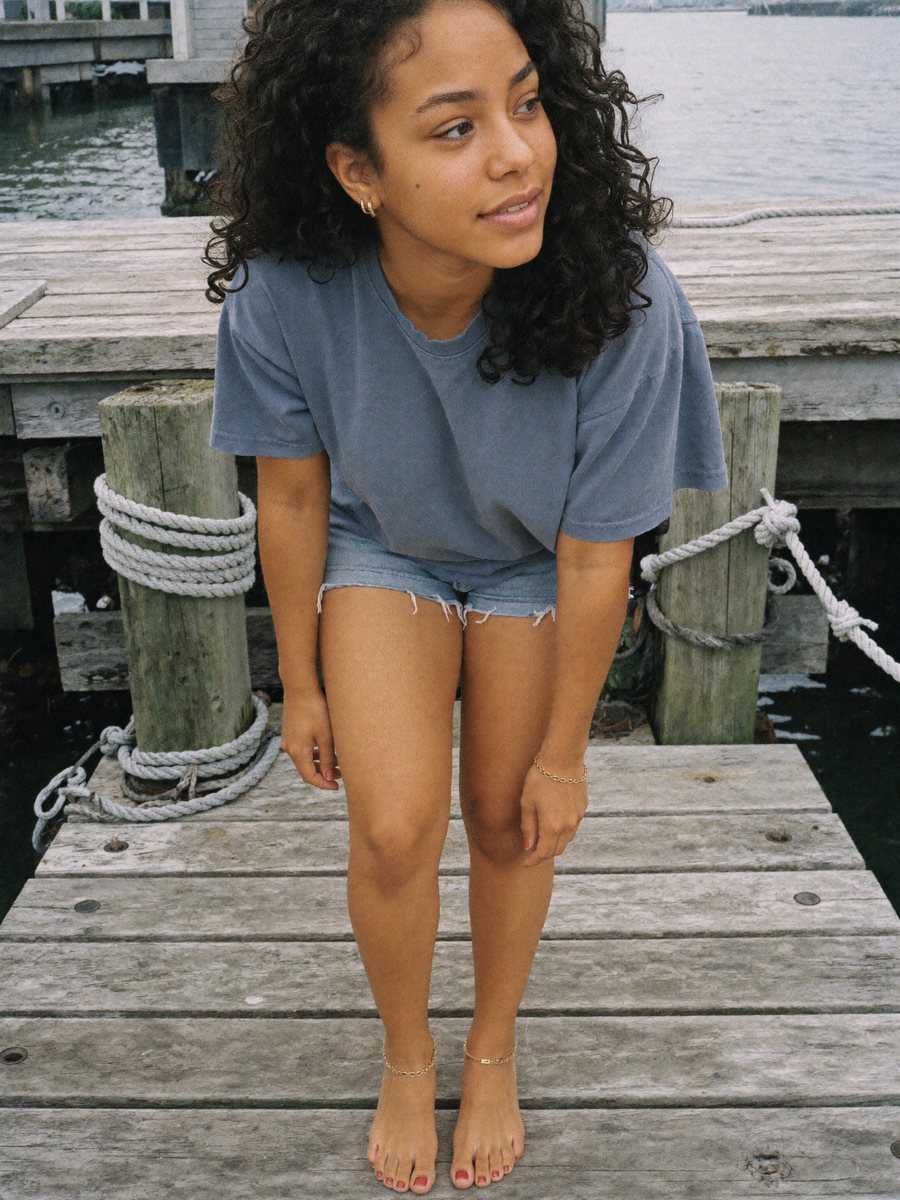 A woman sitting on a weathered dock in a t-shirt and denim shorts, gold anklet at her ankle